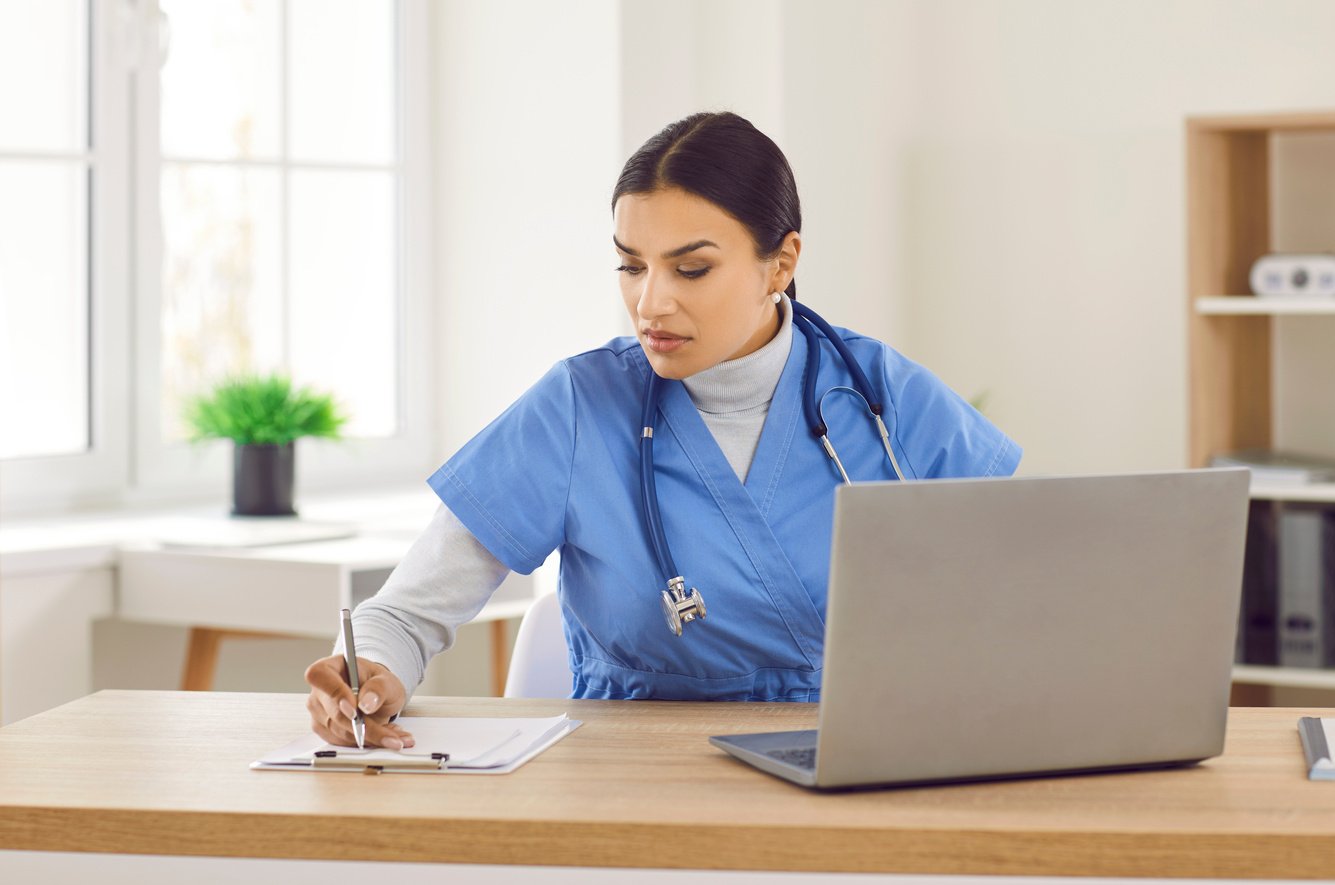 Young Woman Doctor Sitting at Desk, Working on Laptop Computer, and Writing Notes